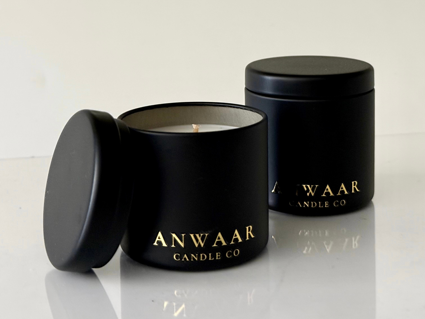 Black candle with open lid and closed container from Anwaar Candle Co on a reflective surface.