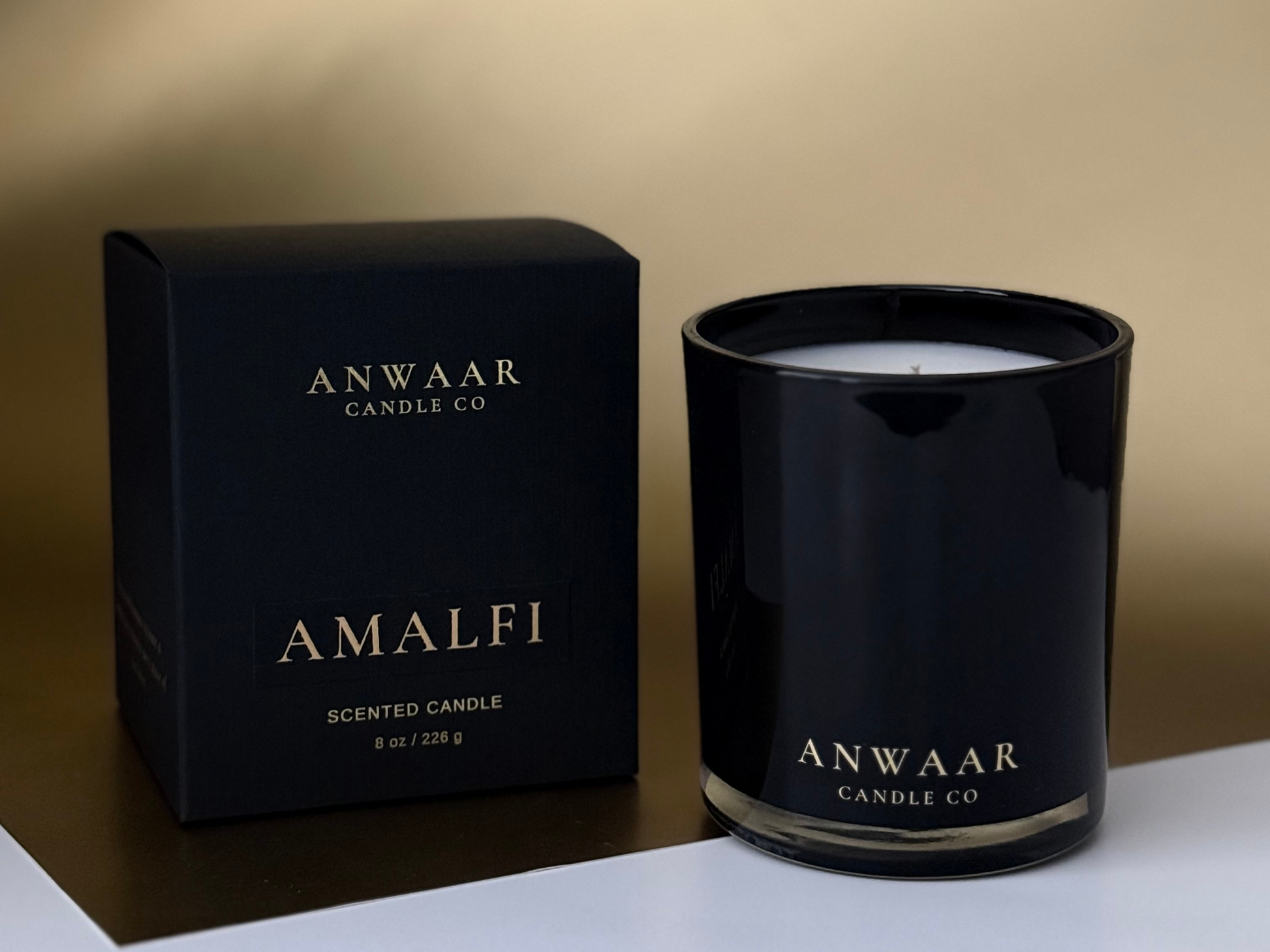 Black candle with 'Anwaar Candle Co' branding next to its packaging on a beige background